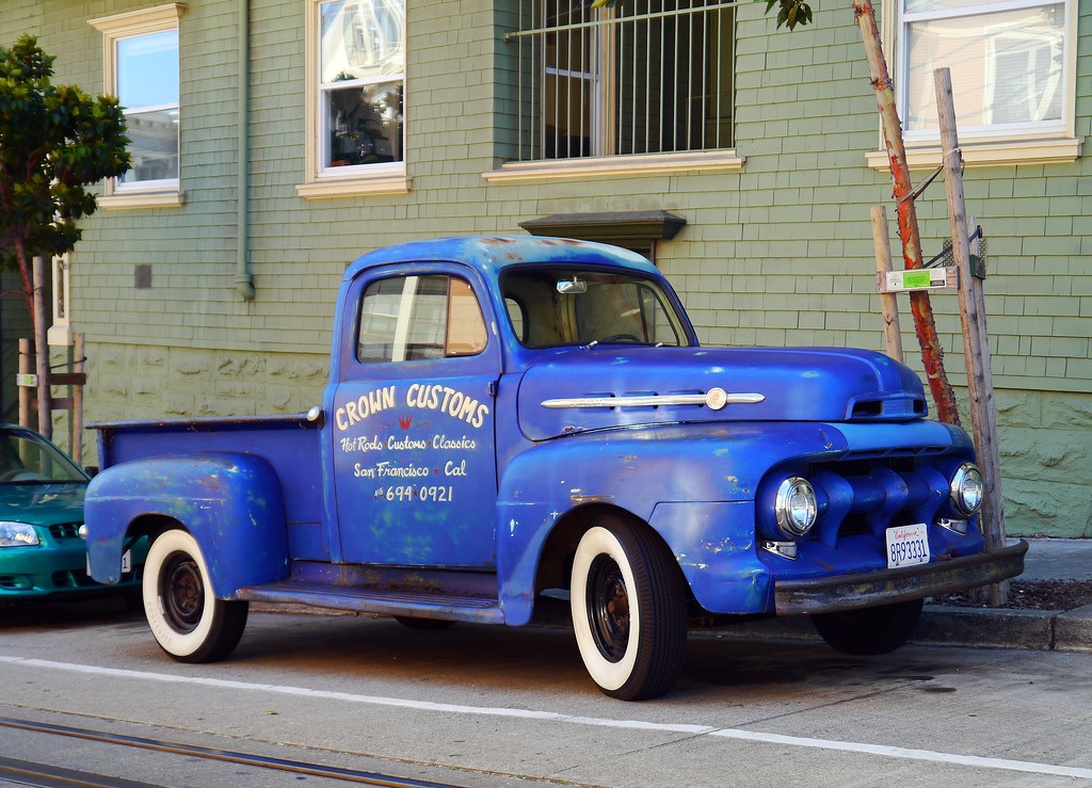 En vackert renoverad klassisk blå Ford pickup med 'CROMN CUSTOMS' på dörren, parkerad på en gata.