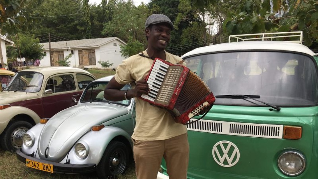 En person med ett dragspel står mellan en klassisk silverfärgad Volkswagen Bubbla och en grön VW-buss.